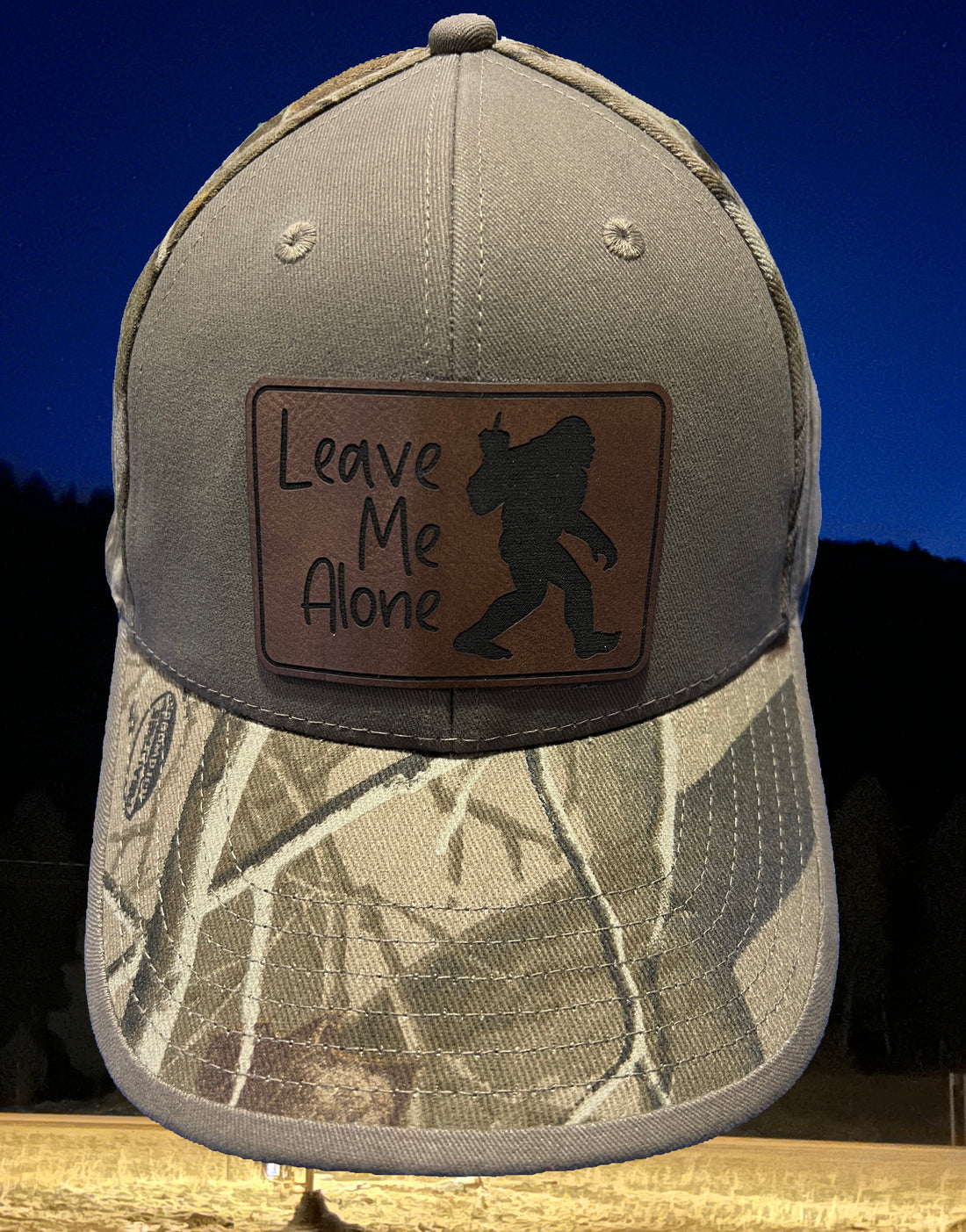 : Image of the "Bigfoot Leave Me Alone" Camo Cap, featuring a camouflage design with the iconic Bigfoot silhouette and humorous message, perfect for outdoor enthusiasts looking to blend in while expressing their need for solitude.